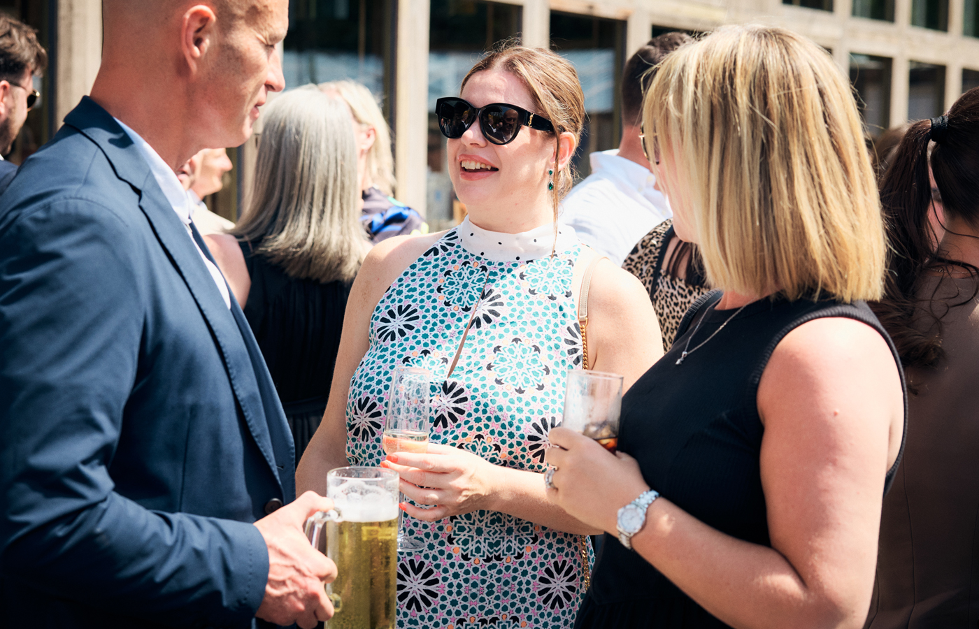 Three professionals in business dress talking and laughing together with drinks at an outdoor industry networking event